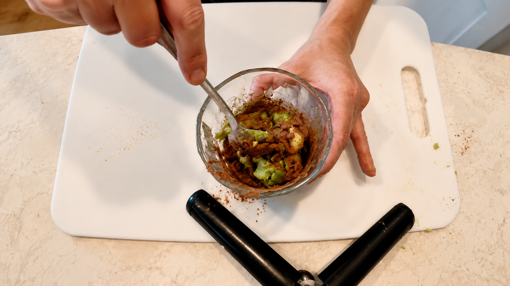 Overhead close-up photo of hands mixing avocado and cocoa powder together in a glass bowl on a white cutting board in a bright kitchen.