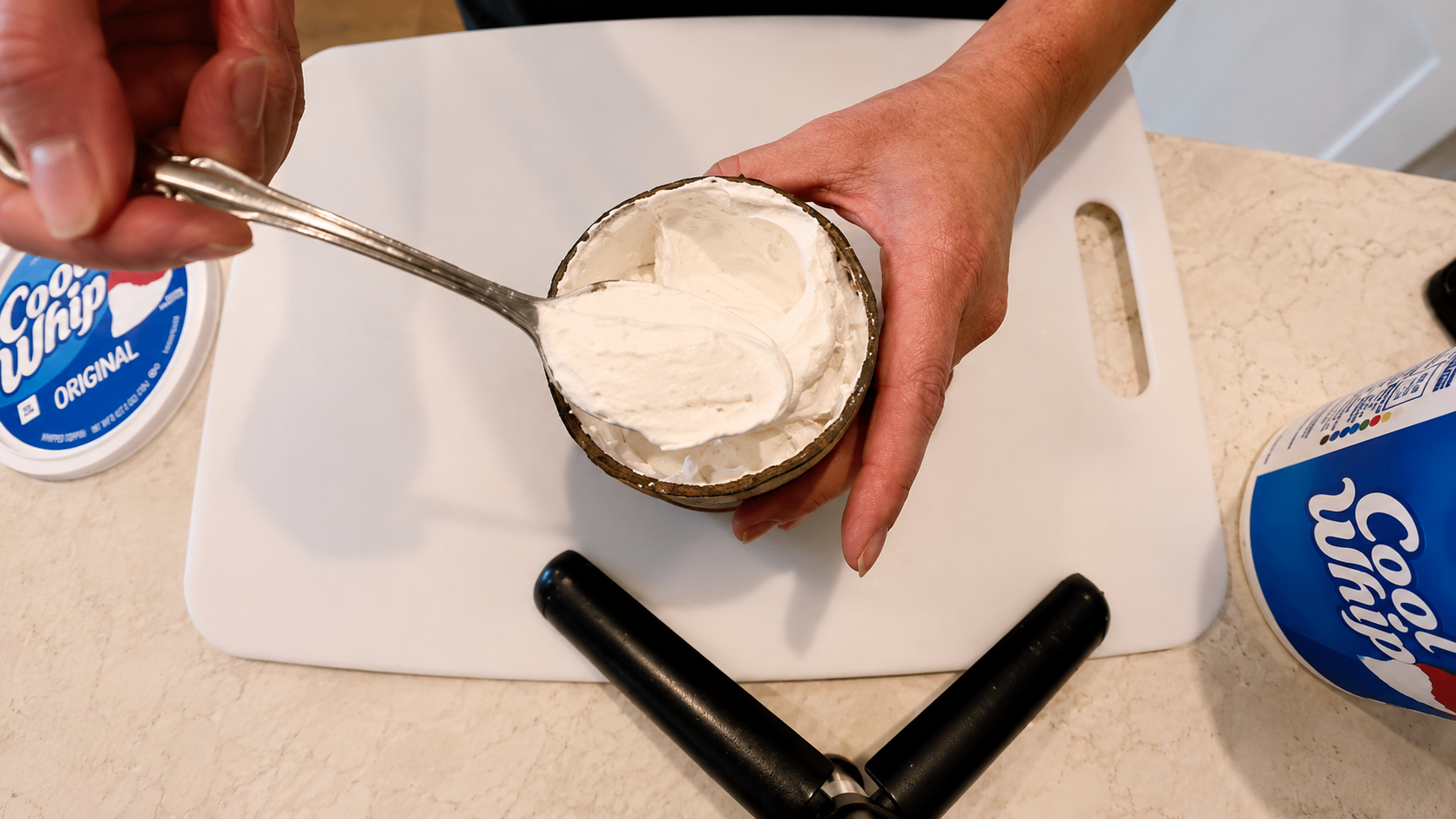 Overhead close-up photo of hands scooping creamy Cool Whip into a dessert bowl on a white cutting board in a bright kitchen.