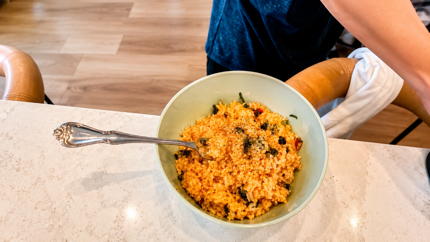 Overhead view of a creamy orange rice bowl topped with seaweed flakes and black pepper on a marble countertop with a silver spoon, photographed in warm natural kitchen lighting.