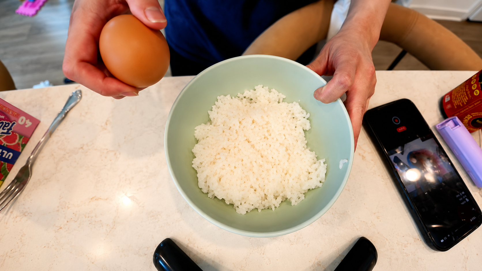 Overhead view of a person holding a brown egg above a pastel green bowl filled with freshly steamed white rice on a marble countertop. A smartphone recording the cooking process and kitchen items sit nearby in a bright cozy kitchen setting.