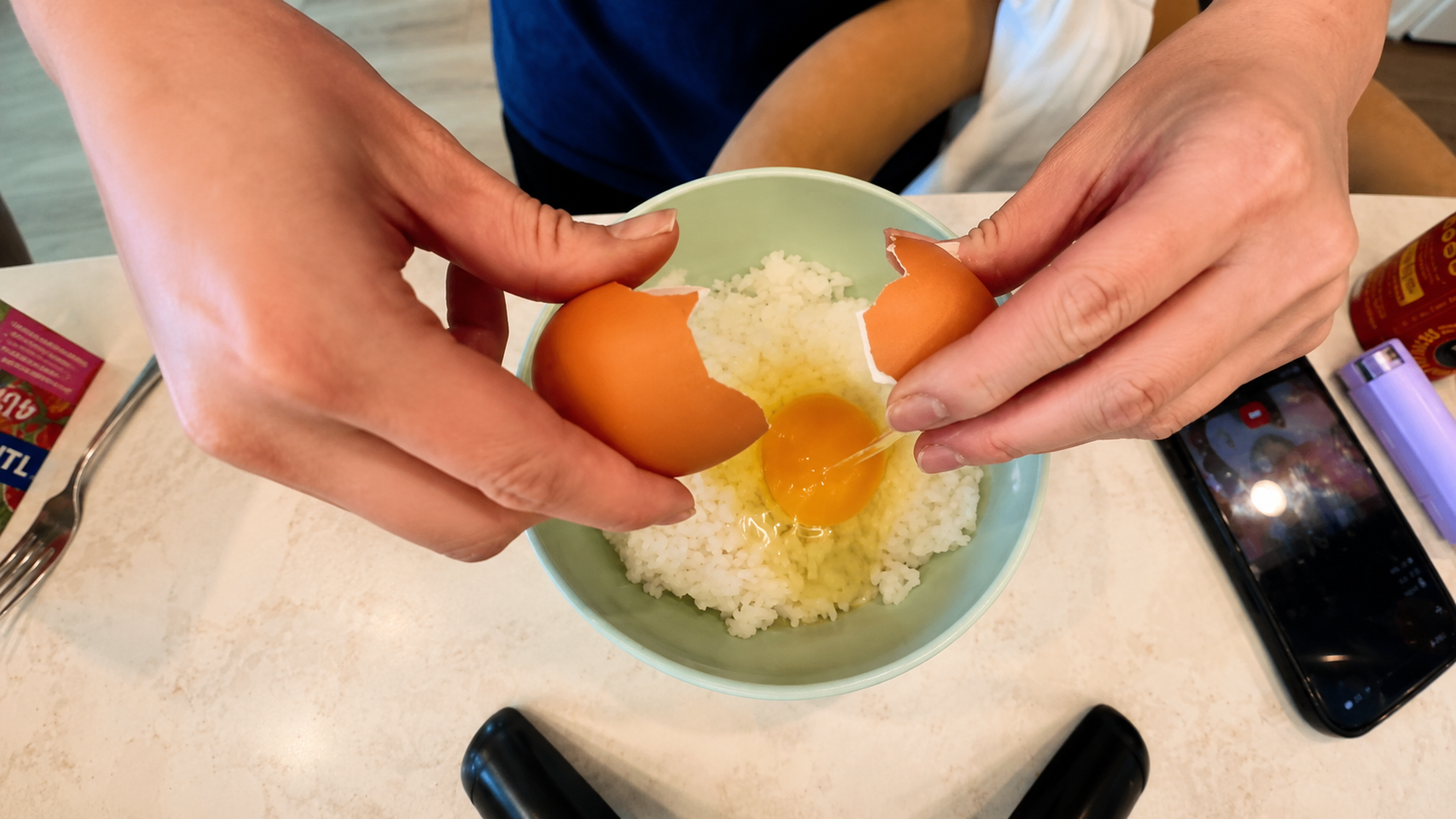 Close up overhead view of a person cracking a raw egg into a pastel green bowl filled with freshly steamed white rice on a marble countertop. A smartphone recording the cooking process and kitchen items sit nearby in a bright cozy kitchen setting.