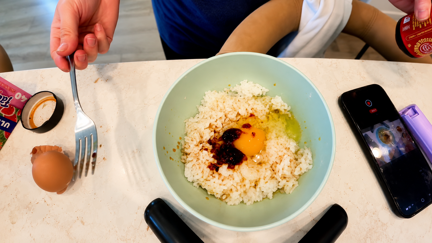 Overhead view of Tamago Kake Gohan in a pastel green bowl with freshly steamed rice, raw egg, and sauce on a marble countertop. A hand holding a fork, cracked eggshell, smartphone recording the cooking process, and kitchen items surround the cozy Japanese inspired rice bowl scene.