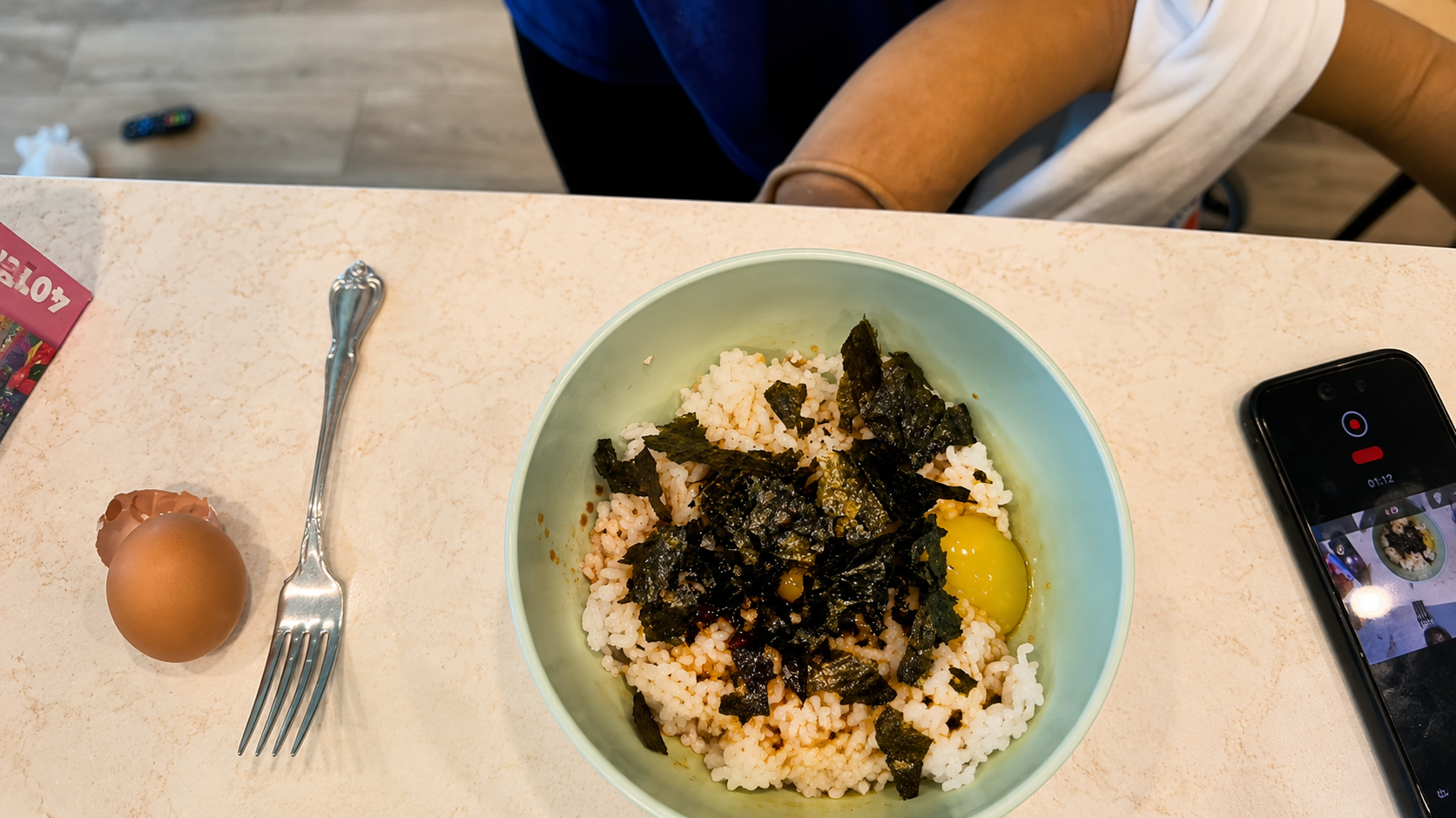 Overhead view of Tamago Kake Gohan in a pastel green bowl topped with seaweed, raw egg, and sauce on a marble countertop. A cracked eggshell, silver fork, smartphone recording the meal, and cozy kitchen details surround the Japanese inspired rice bowl scene.