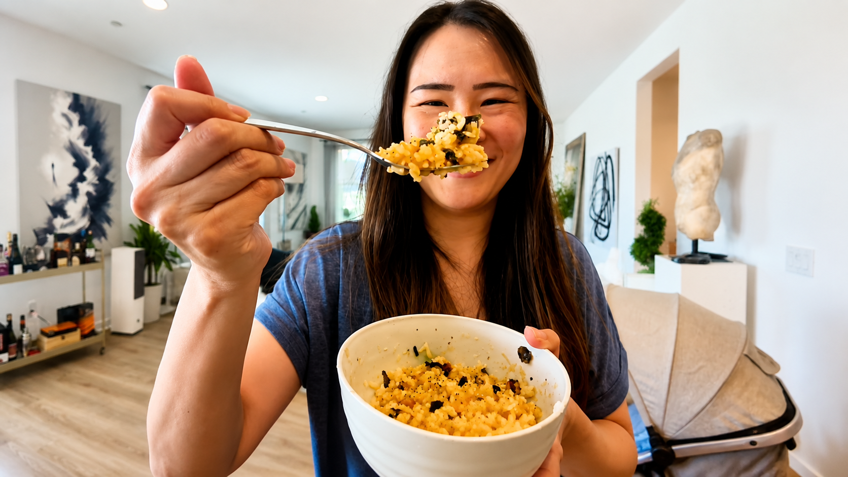 A smiling woman holds a bowl of tamago kake gohan while offering a spoonful toward the camera in a bright modern living room. The rice bowl is filled with creamy seasoned rice topped with seaweed flakes, and the image has a warm professional food photography style with soft natural lighting and a cozy home