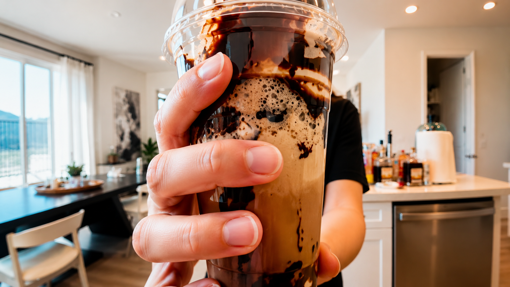 Close-up of a hand holding a crackling iced chocolate latte in a clear plastic cup with rich chocolate swirls, photographed in a bright modern kitchen with soft natural lighting and a professional food photography style.