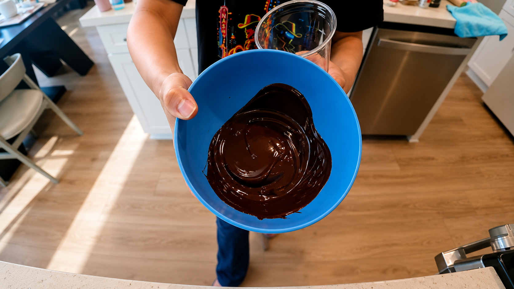 Overhead view of a person holding a blue bowl filled with glossy melted chocolate in a bright modern kitchen, styled with warm lighting and a professional food photography aesthetic.