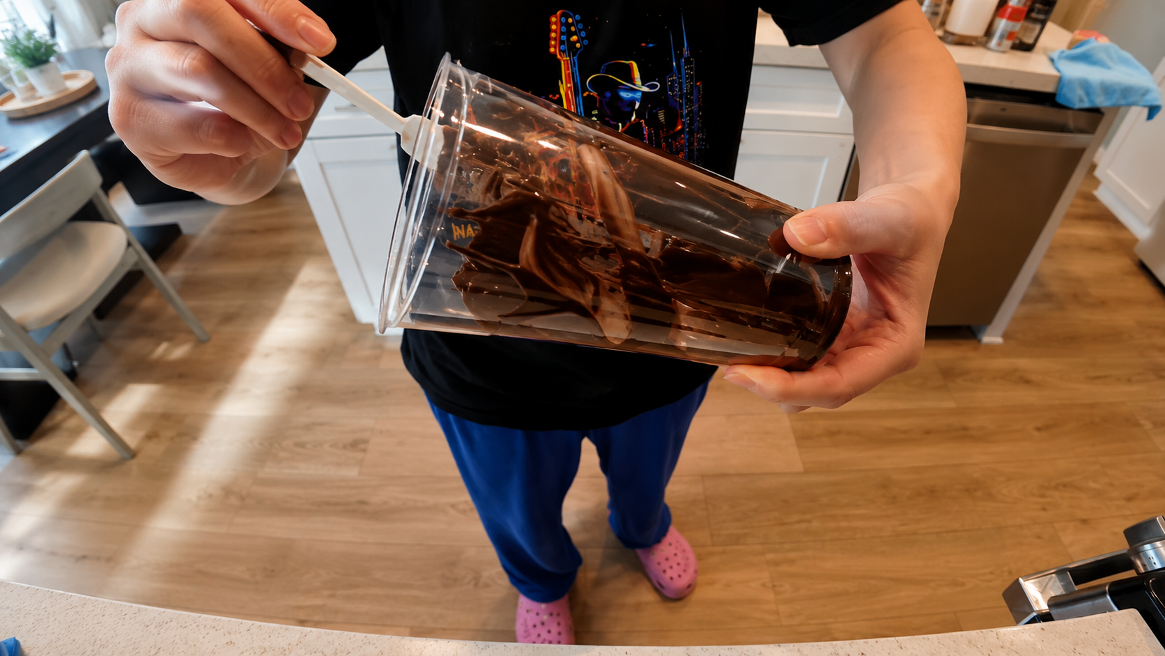 Close-up of a person coating the inside of a clear plastic cup with melted chocolate in a bright modern kitchen, photographed with warm lighting and a professional food photography style.