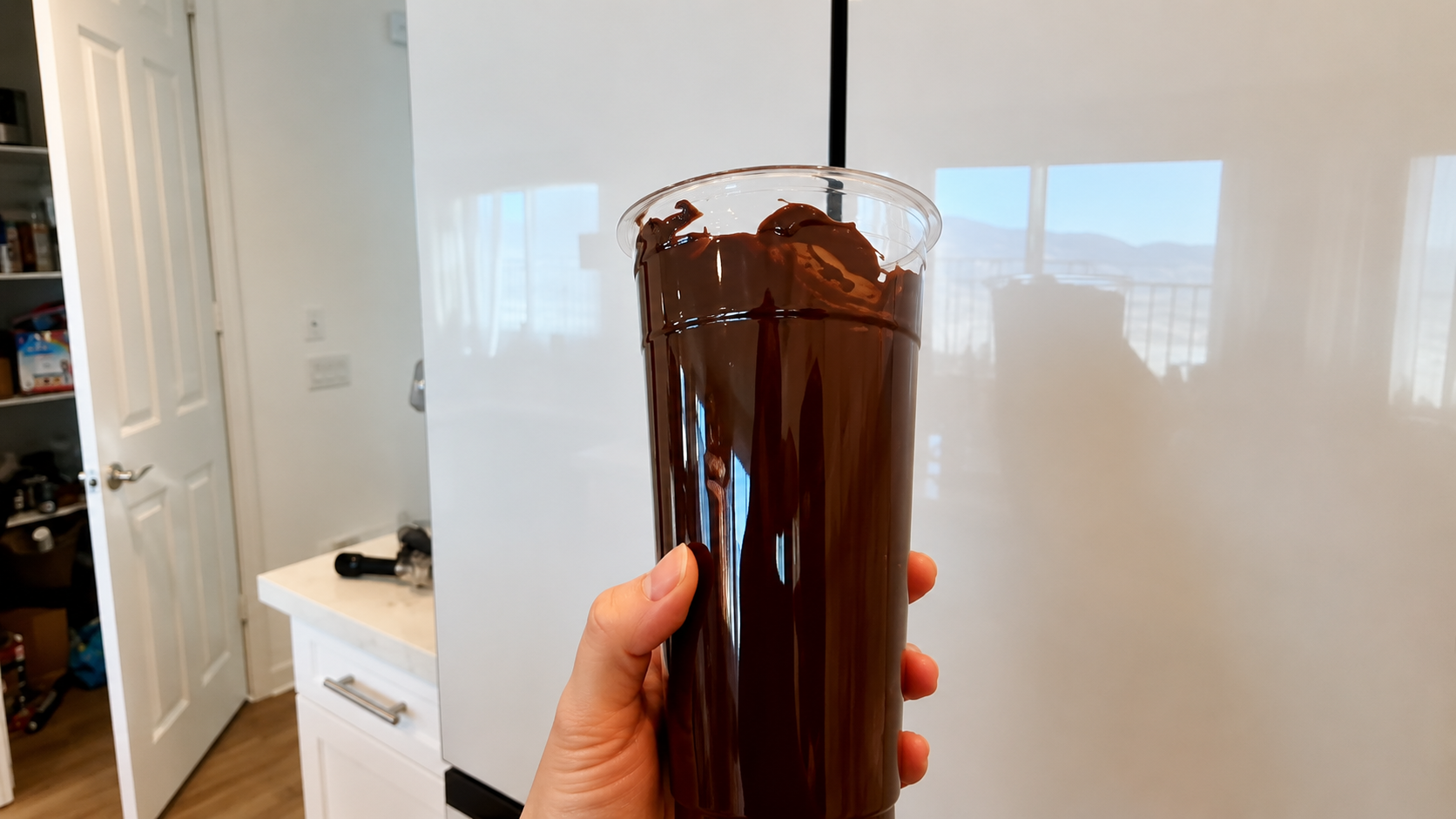 Close-up of a hand holding a tall clear cup coated inside with glossy melted chocolate in a bright modern kitchen with soft natural lighting and a professional food photography aesthetic.