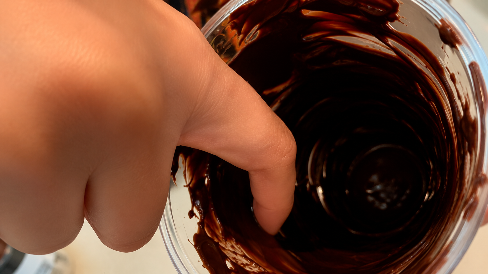 Macro close-up of a hand swirling glossy melted chocolate inside a clear cup, photographed with warm lighting and a rich professional food photography aesthetic.