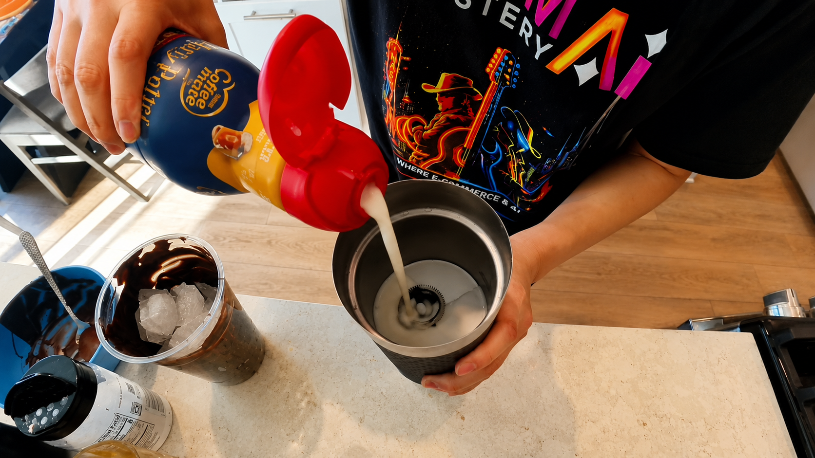 Overhead close-up of a person pouring creamy coffee creamer into a stainless steel frother beside a chocolate-coated iced latte in a bright modern kitchen with a professional food photography aesthetic.
