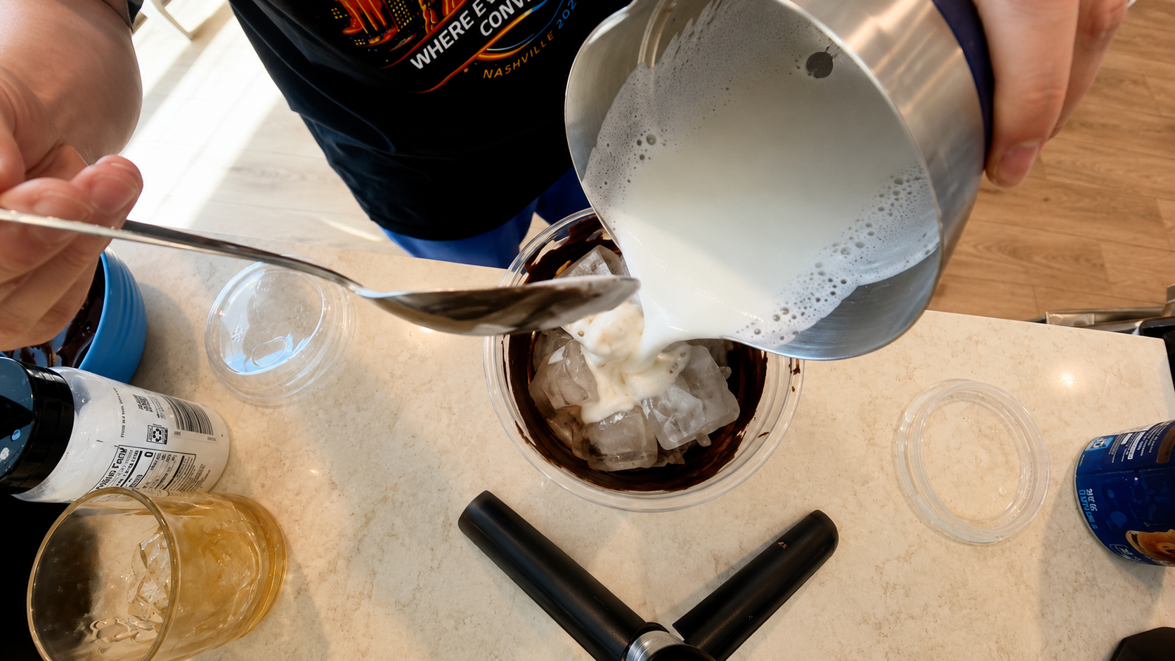Overhead close-up of creamy frothed milk being poured over ice into a chocolate-lined iced latte cup in a bright modern kitchen with a professional food photography aesthetic.