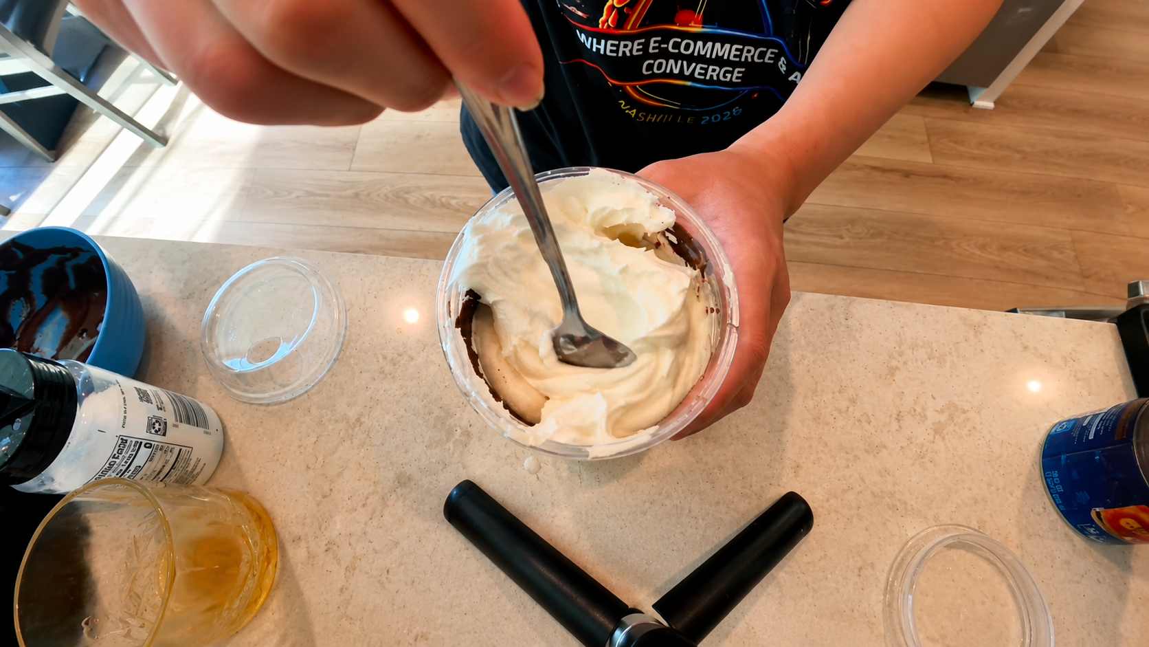 Overhead close-up of a person stirring fluffy whipped topping inside a chocolate-lined iced latte cup on a bright kitchen counter with a professional food photography aesthetic.
