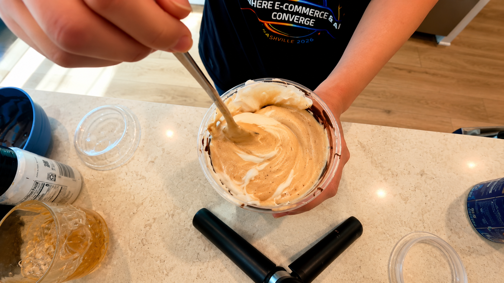 Overhead close-up of a person stirring creamy coffee foam and whipped topping inside a chocolate-lined iced latte cup on a bright kitchen counter with a professional food photography aesthetic.