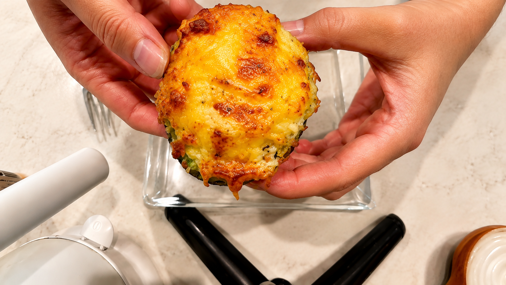 Hands holding a cheesy air fryer avocado egg topped with melted golden cheese over a glass plate on a marble countertop.