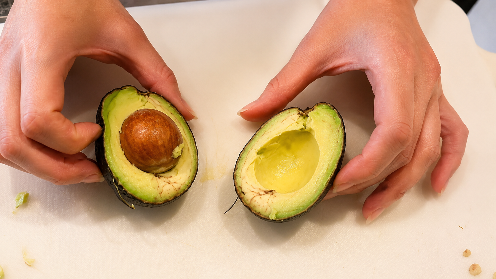 Hands holding two fresh avocado halves on a white cutting board, with one half containing the avocado pit and the other showing the creamy center removed.