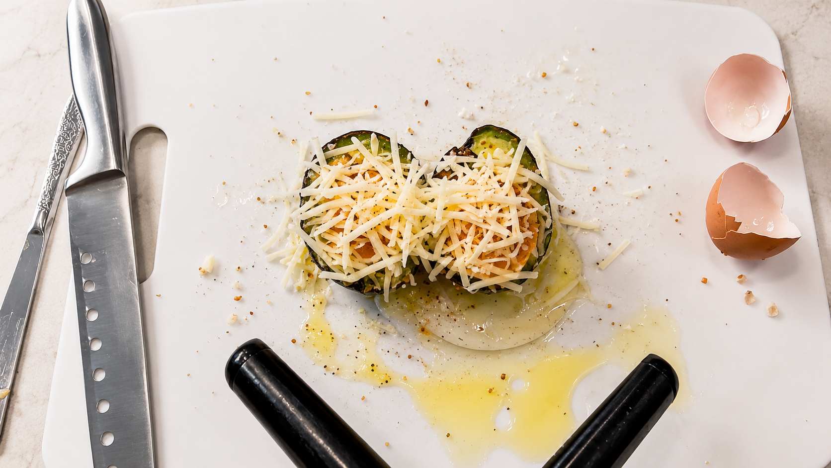 Overhead food photography shot of two avocado halves filled with egg and topped generously with shredded mozzarella cheese on a white cutting board. The scene features glossy olive oil, scattered seasoning, cracked eggshells, and stainless steel kitchen knives on a light marble countertop, creating a bright and professional cooking prep aesthetic.