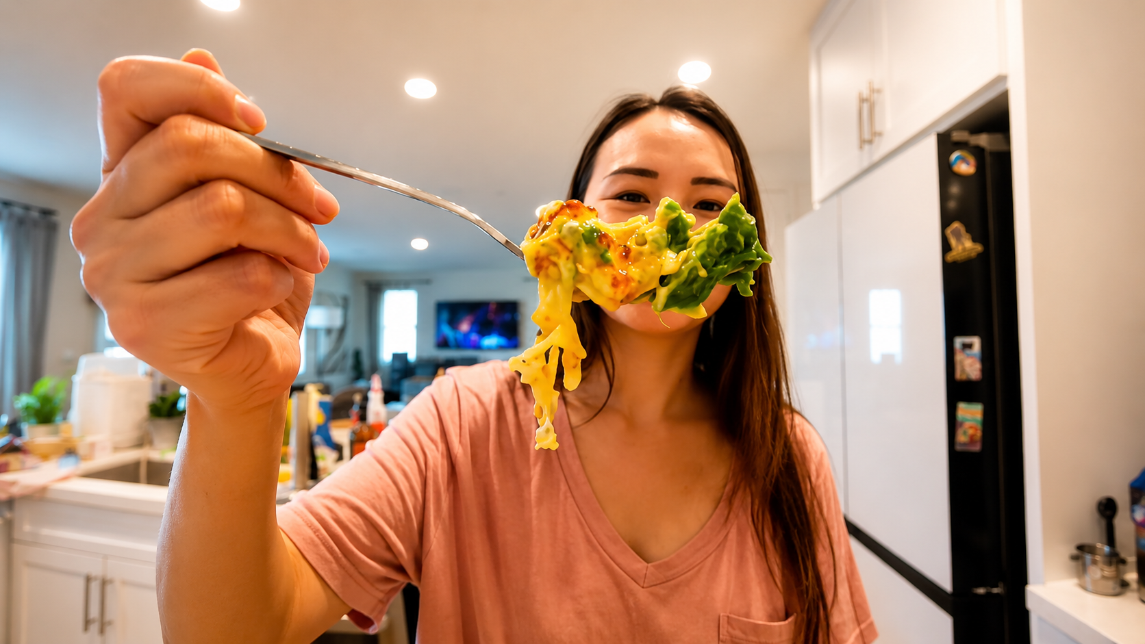A woman in a pink shirt holds up a forkful of cheesy avocado egg in a modern kitchen. Melted golden cheese stretches from the fork while creamy avocado and fluffy egg create a rich, indulgent bite. The softly blurred kitchen background and warm lighting give the image a cozy, professional food photography feel.