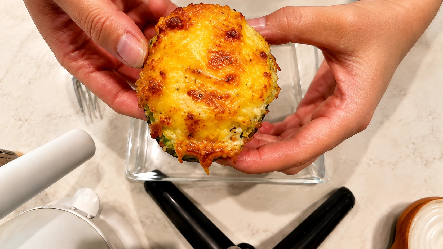 Hands holding a cheesy air fryer avocado egg topped with melted golden cheese over a glass plate on a marble countertop.