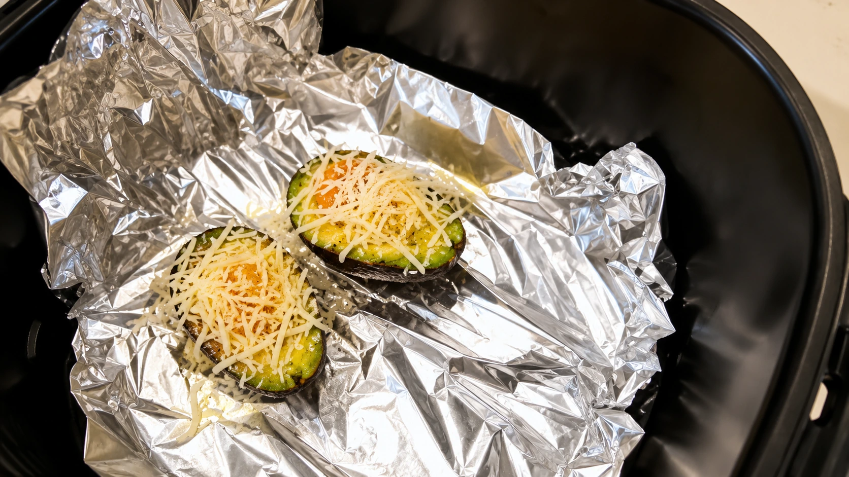 Close-up overhead shot of an air fryer basket lined with crinkled aluminum foil holding two avocado halves topped with shredded cheese and egg filling. The glossy foil reflects warm kitchen lighting, while the vibrant green avocado and melted cheese create a rich, appetizing food photography scene.