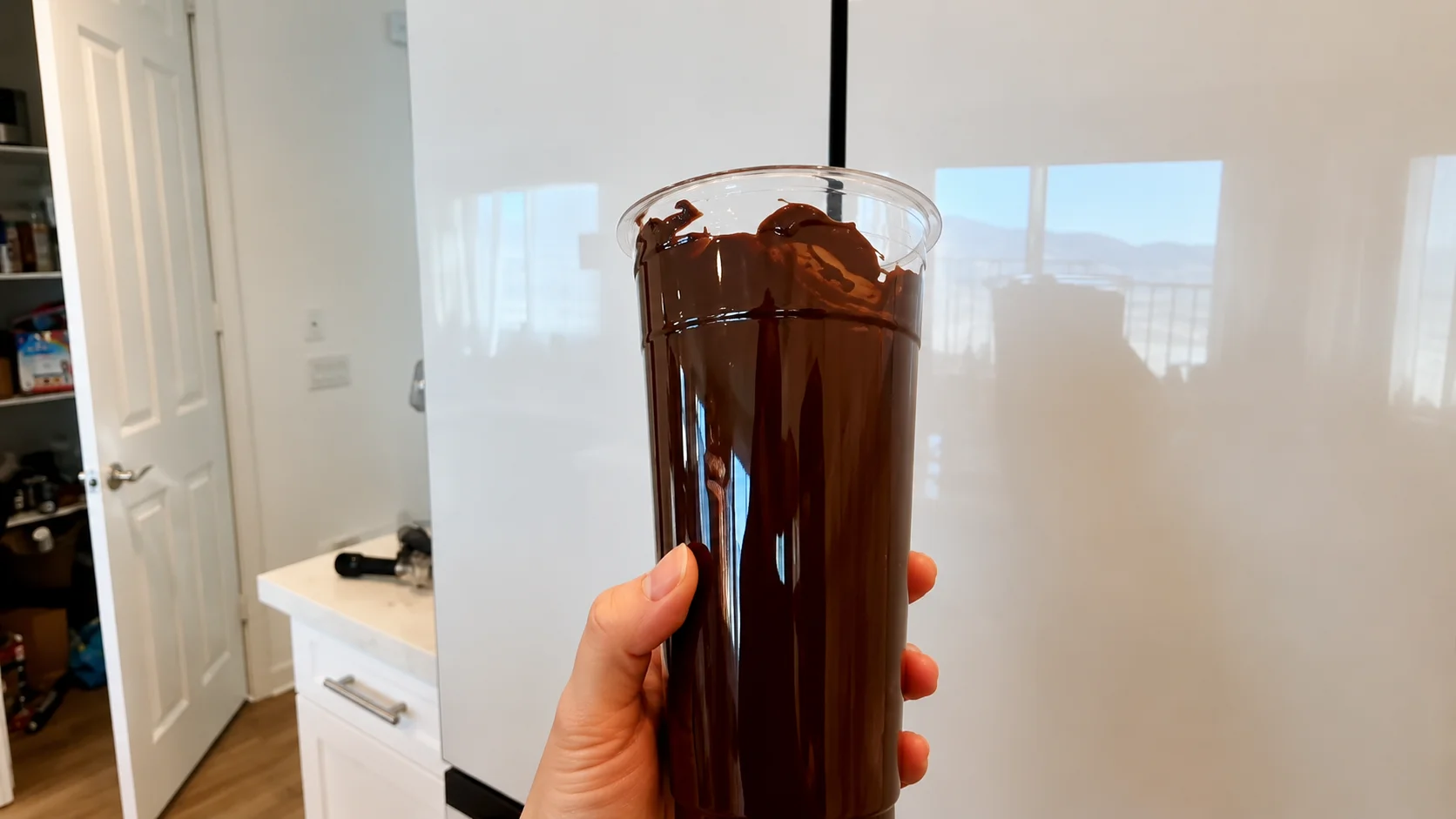 Close-up of a hand holding a tall clear cup coated inside with glossy melted chocolate in a bright modern kitchen with soft natural lighting and a professional food photography aesthetic.