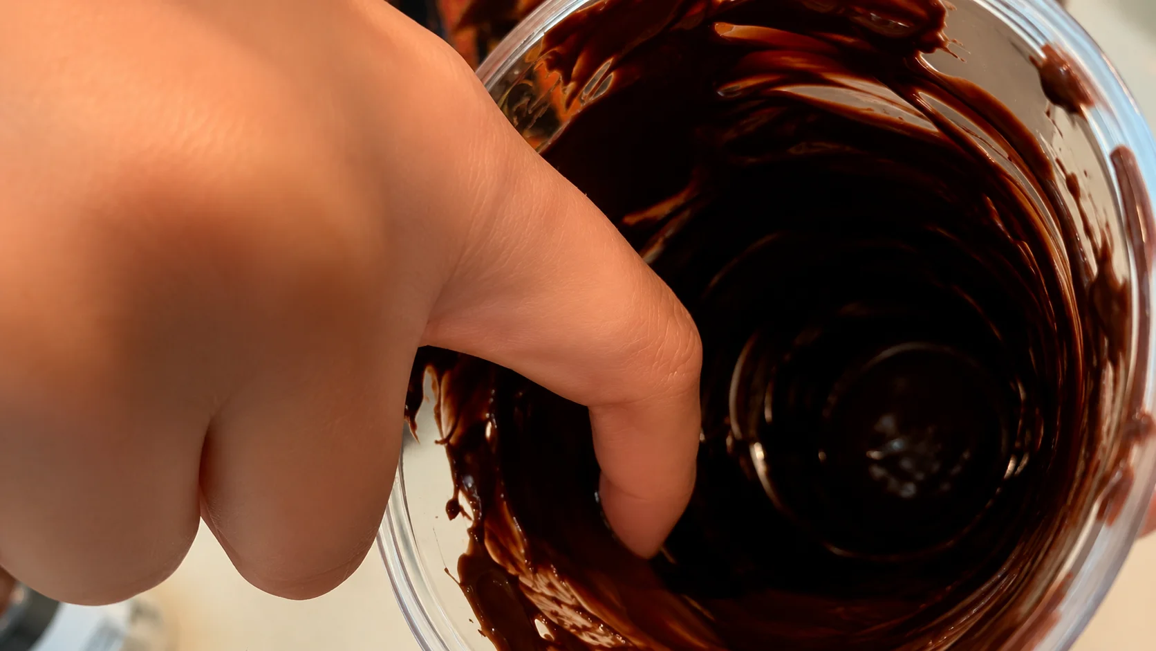Macro close-up of a hand swirling glossy melted chocolate inside a clear cup, photographed with warm lighting and a rich professional food photography aesthetic.
