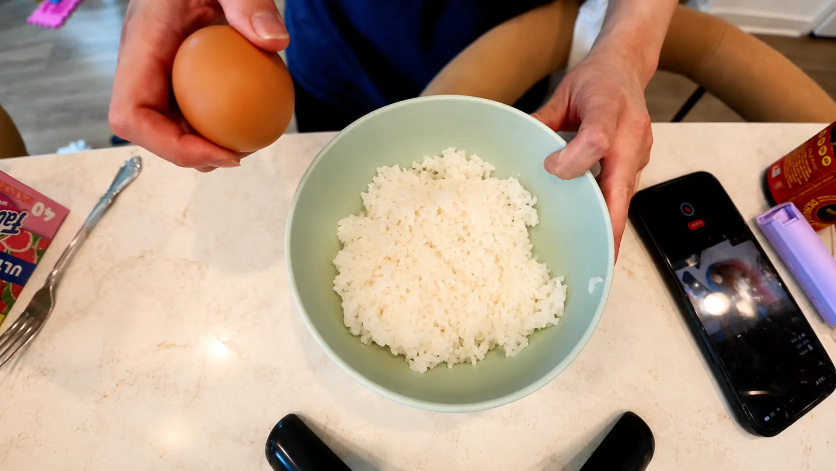 Overhead view of a person holding a brown egg above a pastel green bowl filled with freshly steamed white rice on a marble countertop. A smartphone recording the cooking process and kitchen items sit nearby in a bright cozy kitchen setting.