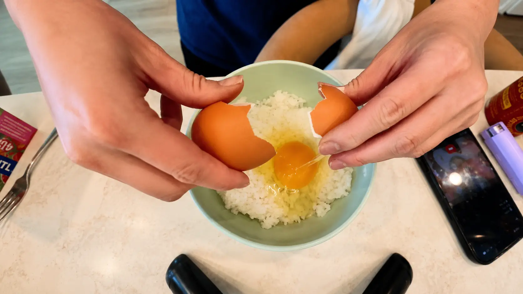 Close up overhead view of a person cracking a raw egg into a pastel green bowl filled with freshly steamed white rice on a marble countertop. A smartphone recording the cooking process and kitchen items sit nearby in a bright cozy kitchen setting.