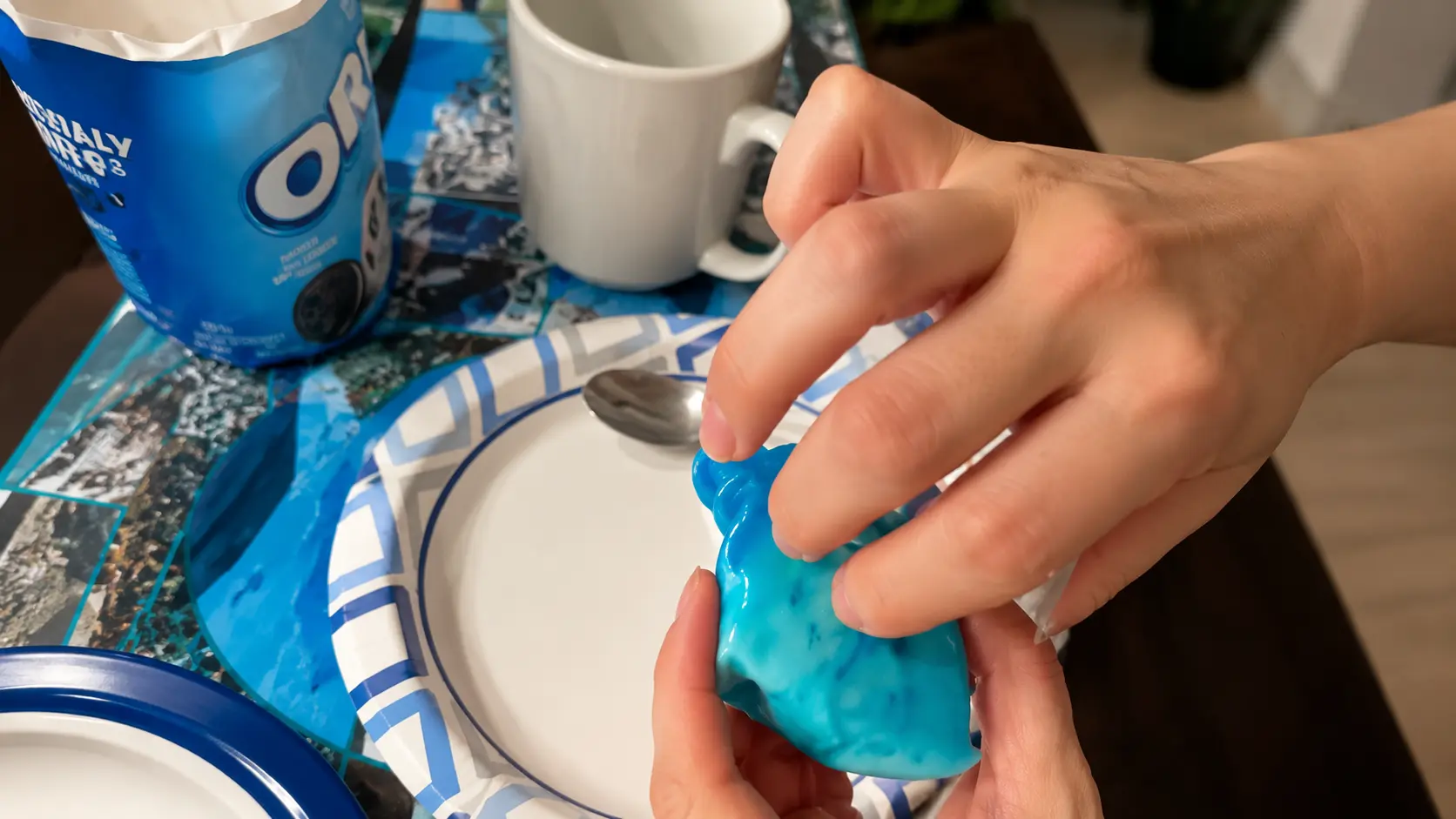 Close-up of hands stretching and squeezing a bright blue Oreo-inspired dessert or snack over a white paper plate on a tabletop. An open Oreo package and a white mug sit in the background, softly blurred. The image is brightly lit with a shallow depth of field, highlighting the glossy texture of the blue treat and creating a professional food photography appearance.