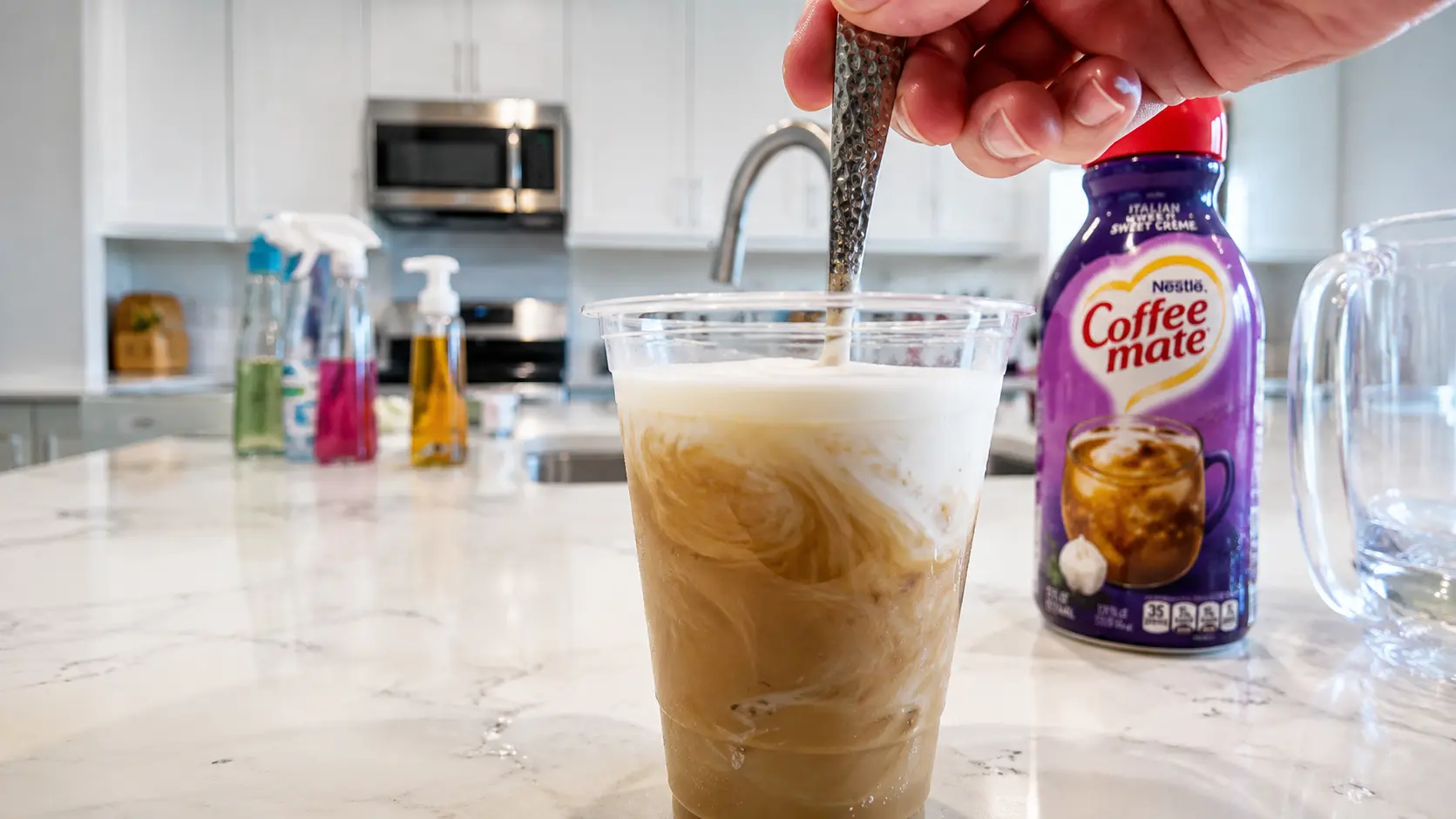 A close-up of a creamy iced coffee being stirred in a clear plastic cup on a white marble countertop. Swirls of coffee and cream create a rich marbled effect beneath a layer of frothy foam. A hand holds a textured metal spoon while mixing the drink. In the softly blurred background, a bottle of Coffee Mate Italian Sweet Crème creamer sits beside a bright, modern kitchen with white cabinets, stainless steel appliances, and natural daylight streaming through the space. The image has a clean, professional food photography style with sharp focus on the beverage and a shallow depth of field.