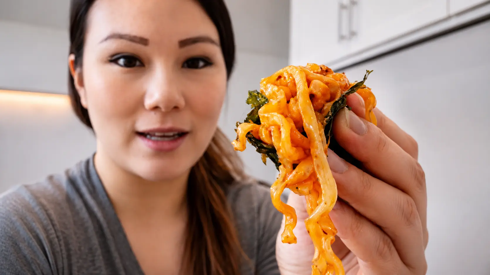 Woman holding cheesy Buldak Carbonara ramen wrapped in roasted seaweed, showing glossy spicy noodles close to camera.