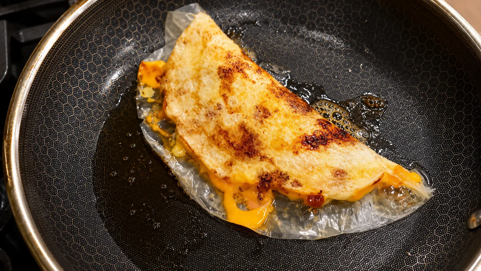 Close-up of a crispy Vietnamese rice paper pizza cooking in a nonstick frying pan. The folded rice paper is golden brown with toasted spots and filled with melted cheese that oozes from the edges. Hot oil glistens around the pizza, highlighting its crunchy texture and cheesy center. The overhead angle and professional food photography style emphasize the rich colors, crisp surface, and irresistible gooey cheese.