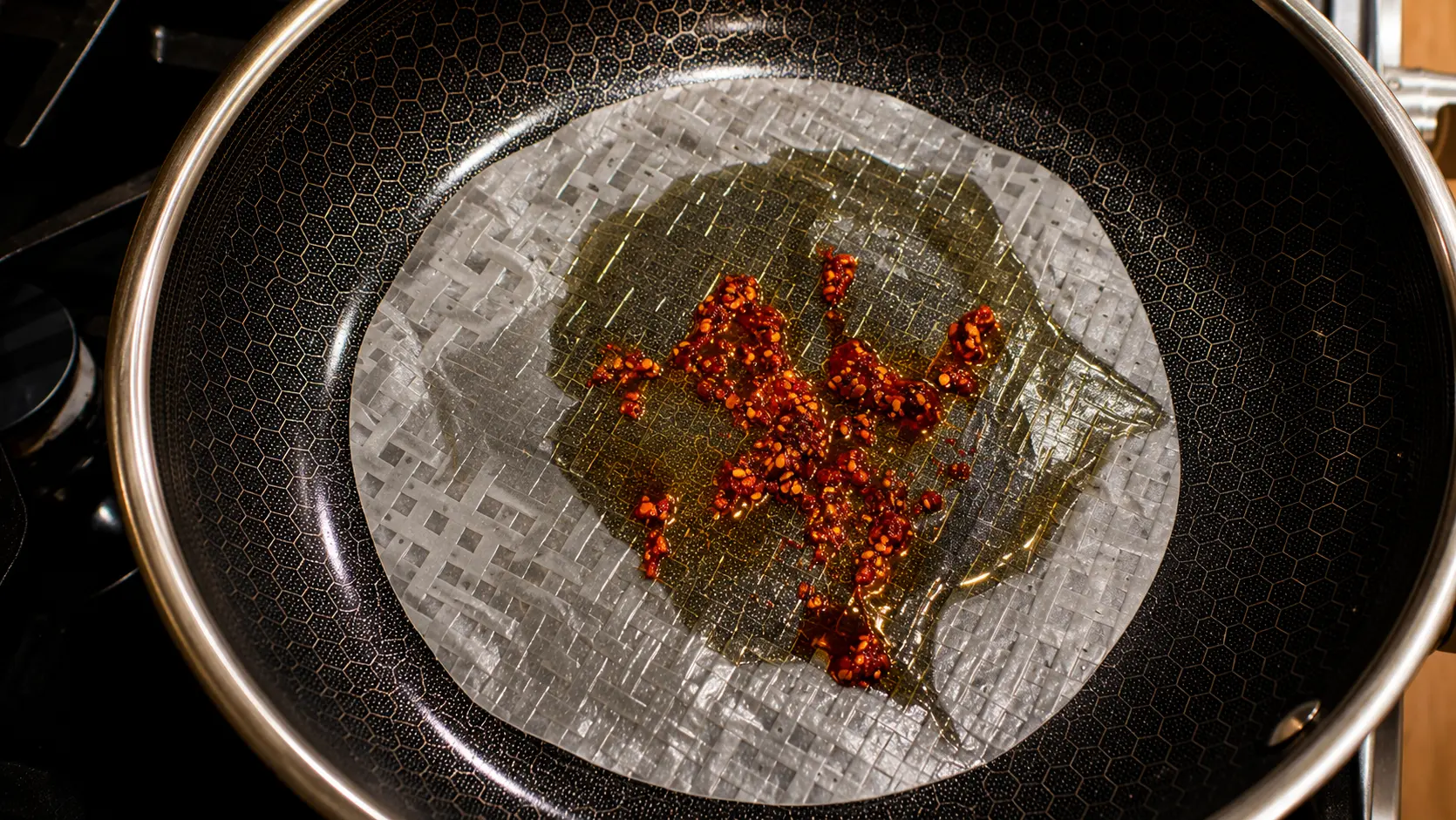 Close-up overhead view of a Vietnamese rice paper pizza in progress, cooking in a nonstick skillet with a hexagon-patterned surface. A translucent rice paper sheet is coated with glossy chili oil and sprinkled with vibrant red chili flakes. The oil glistens under warm lighting, highlighting the delicate texture of the rice paper and creating an appetizing contrast against the dark pan. The professional food photography style emphasizes the rich colors, crisp details, and savory ingredients before the pizza is assembled.
