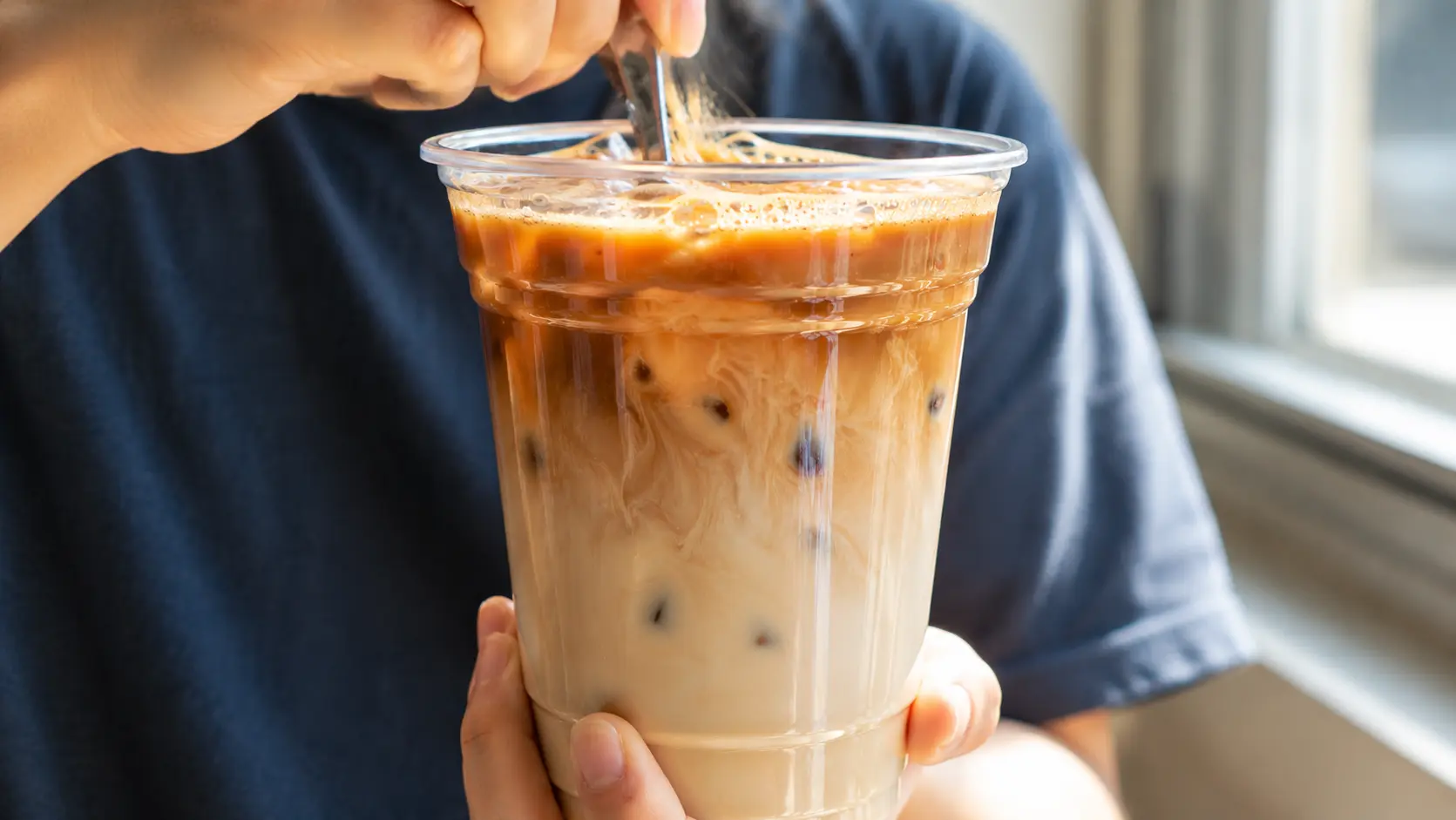 Hyper-realistic Dalgona iced coffee in a clear plastic cup on a white marble countertop, featuring creamy milk swirled with rich espresso and topped with thick whipped coffee foam in a modern kitchen setting.