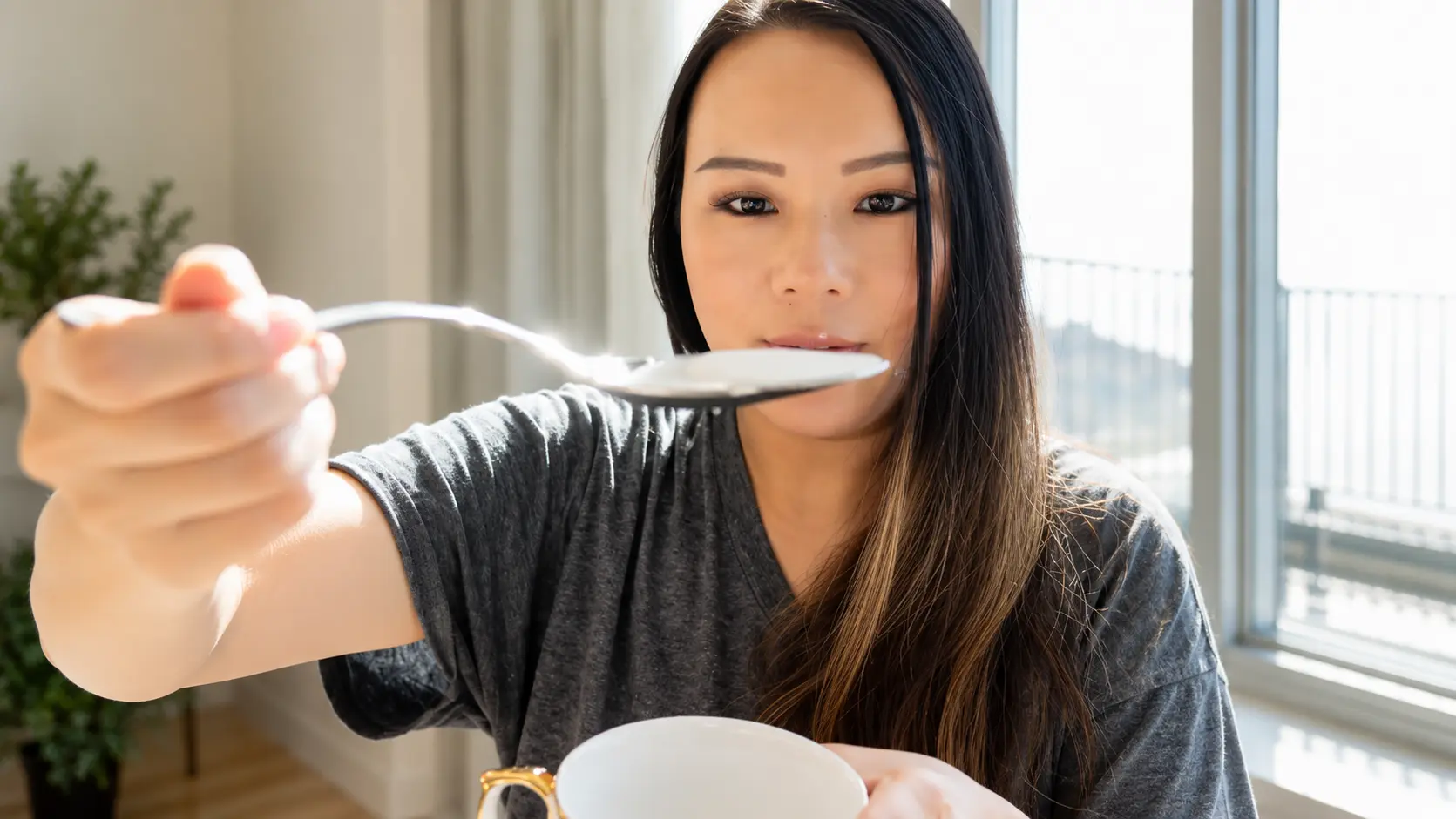 Close-up of a woman holding a spoonful of granulated white sugar toward the camera in a bright kitchen, with the sugar crystals sharply focused and clearly visible.