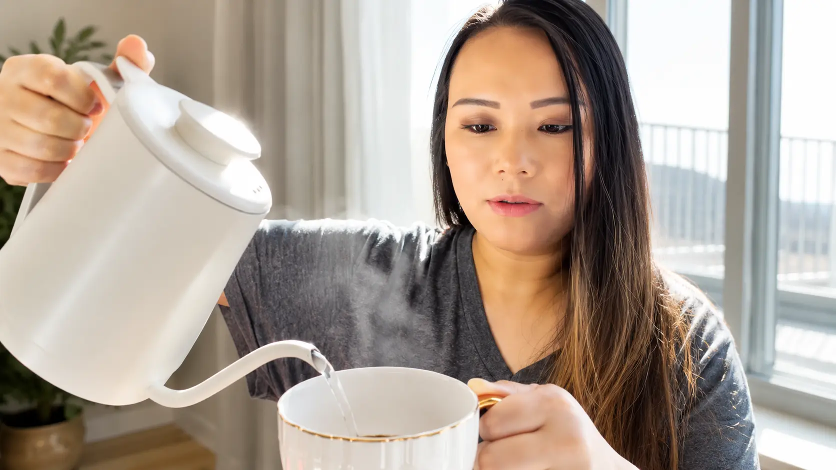 Woman pouring steaming hot water from a white gooseneck kettle into a white ceramic cup of dark coffee, with visible water stream and rising steam, softly lit by natural window light in a cozy kitchen setting.