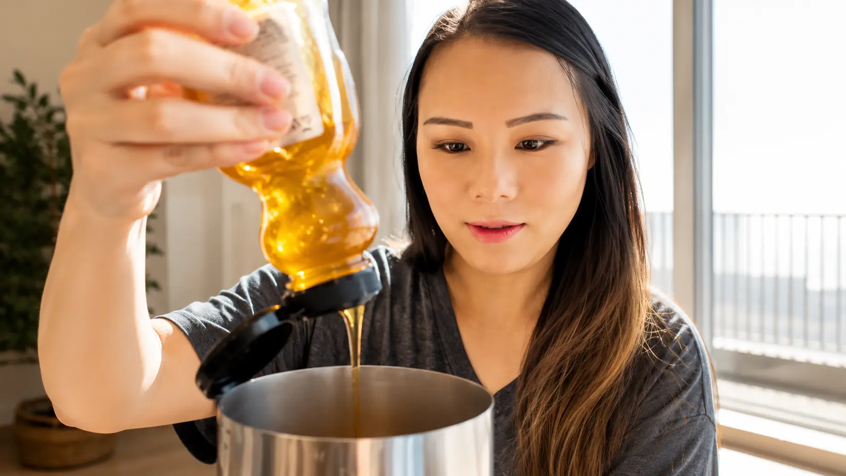 Woman drizzling golden honey from a squeeze bottle into a stainless steel milk frother filled with coffee, with visible honey stream and soft natural window light creating a warm, cozy kitchen atmosphere.