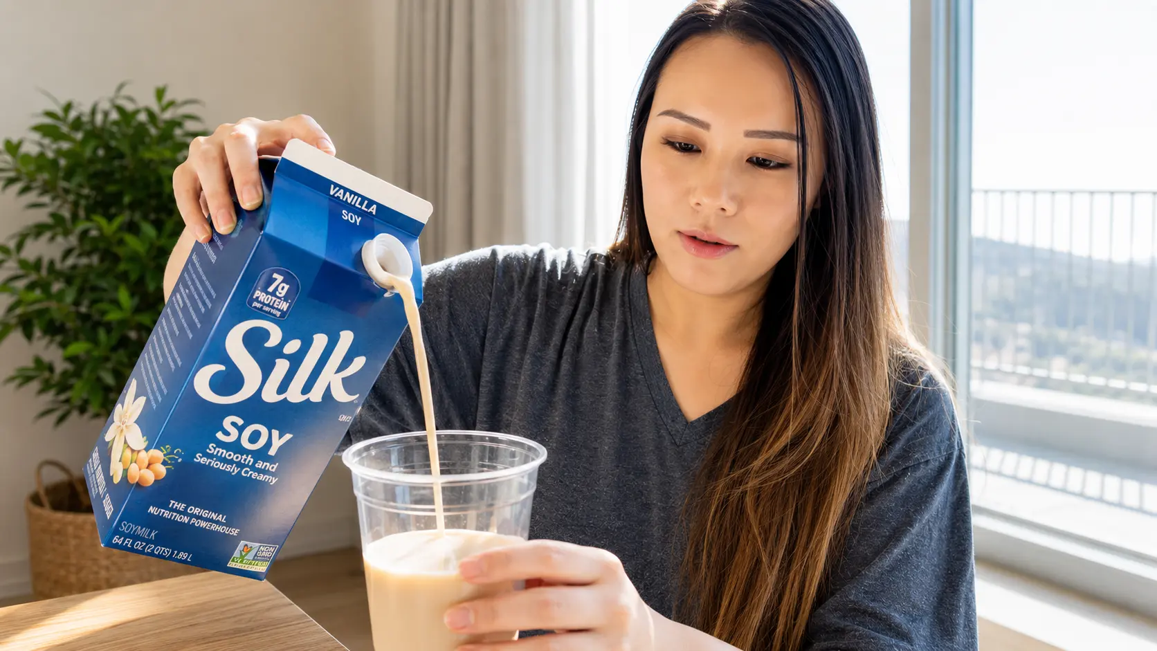 Woman pouring soy milk from a blue carton into a clear plastic cup filled with iced coffee, with visible milk stream and ice cubes, softly lit by natural window light in a cozy kitchen.