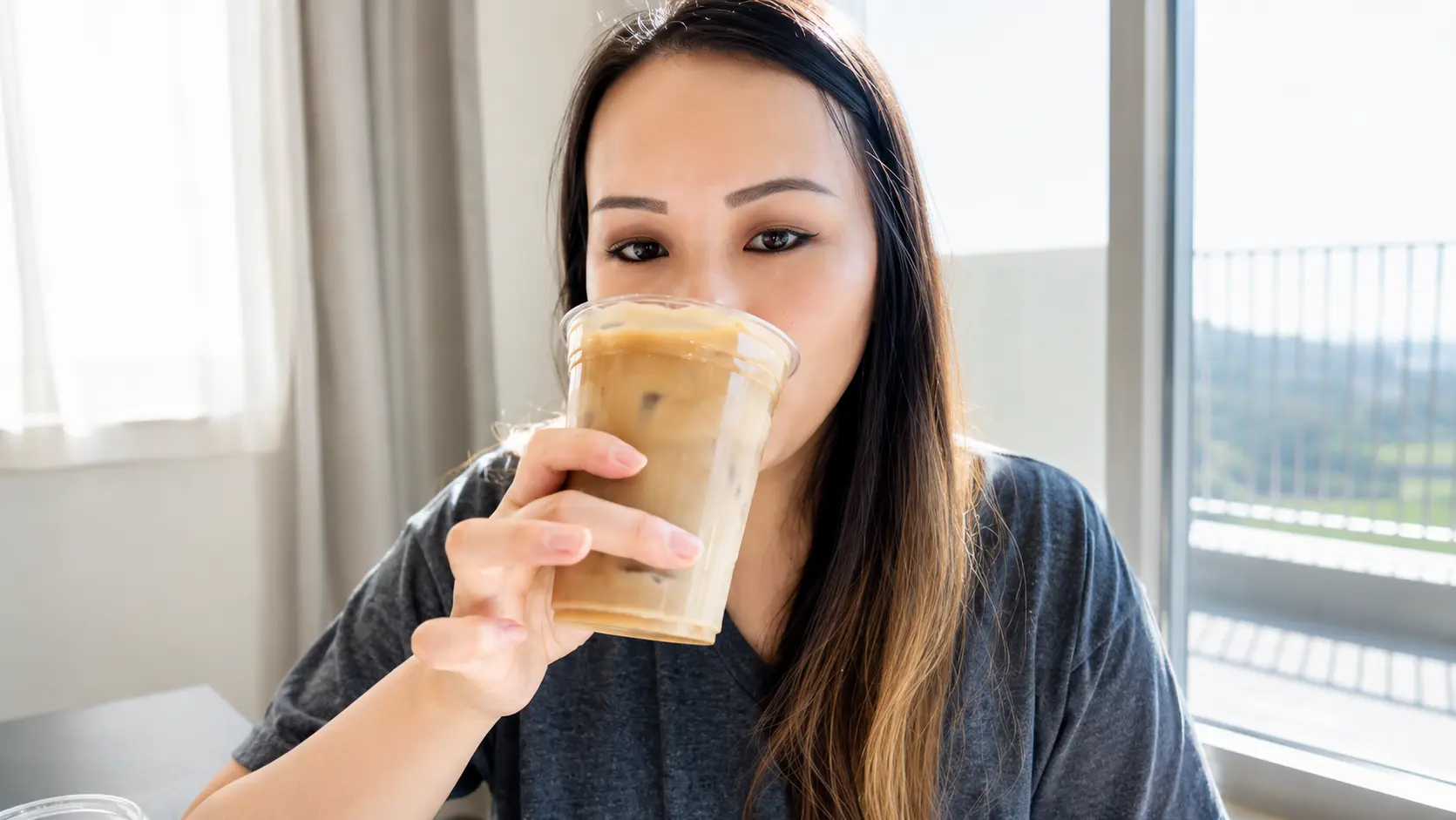 Woman sipping iced dalgona coffee from a clear plastic cup topped with cinnamon-dusted whipped coffee foam, with layered milk and ice visible in soft natural window light.
