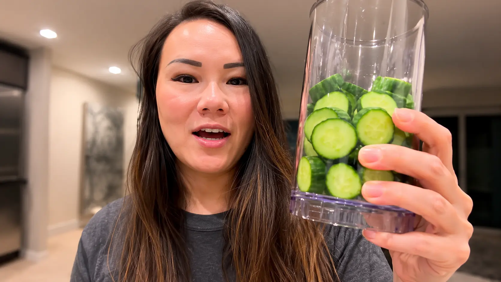 Close-up of a woman holding a clear glass filled with freshly sliced English cucumbers, the vibrant green rounds in sharp focus with visible seeds and crisp texture, set against a softly blurred modern kitchen background.