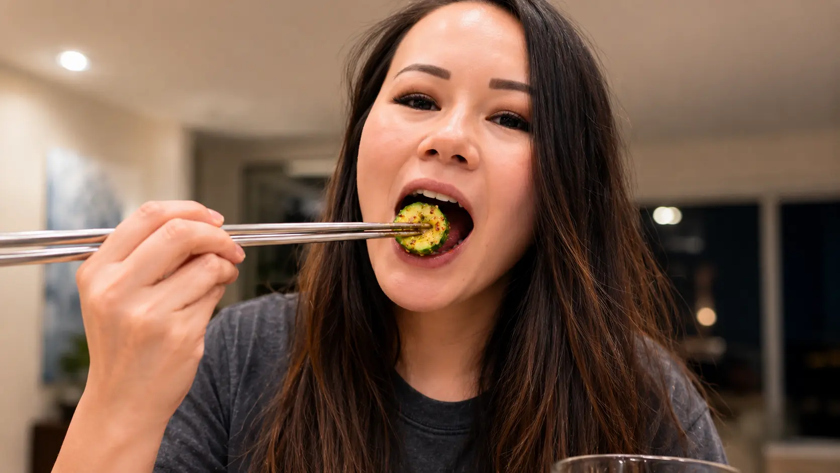 Close-up of a woman eating a marinated cucumber slice with chopsticks, the glossy cucumber coated in sesame seeds and chili flakes in sharp focus against a softly blurred kitchen background.