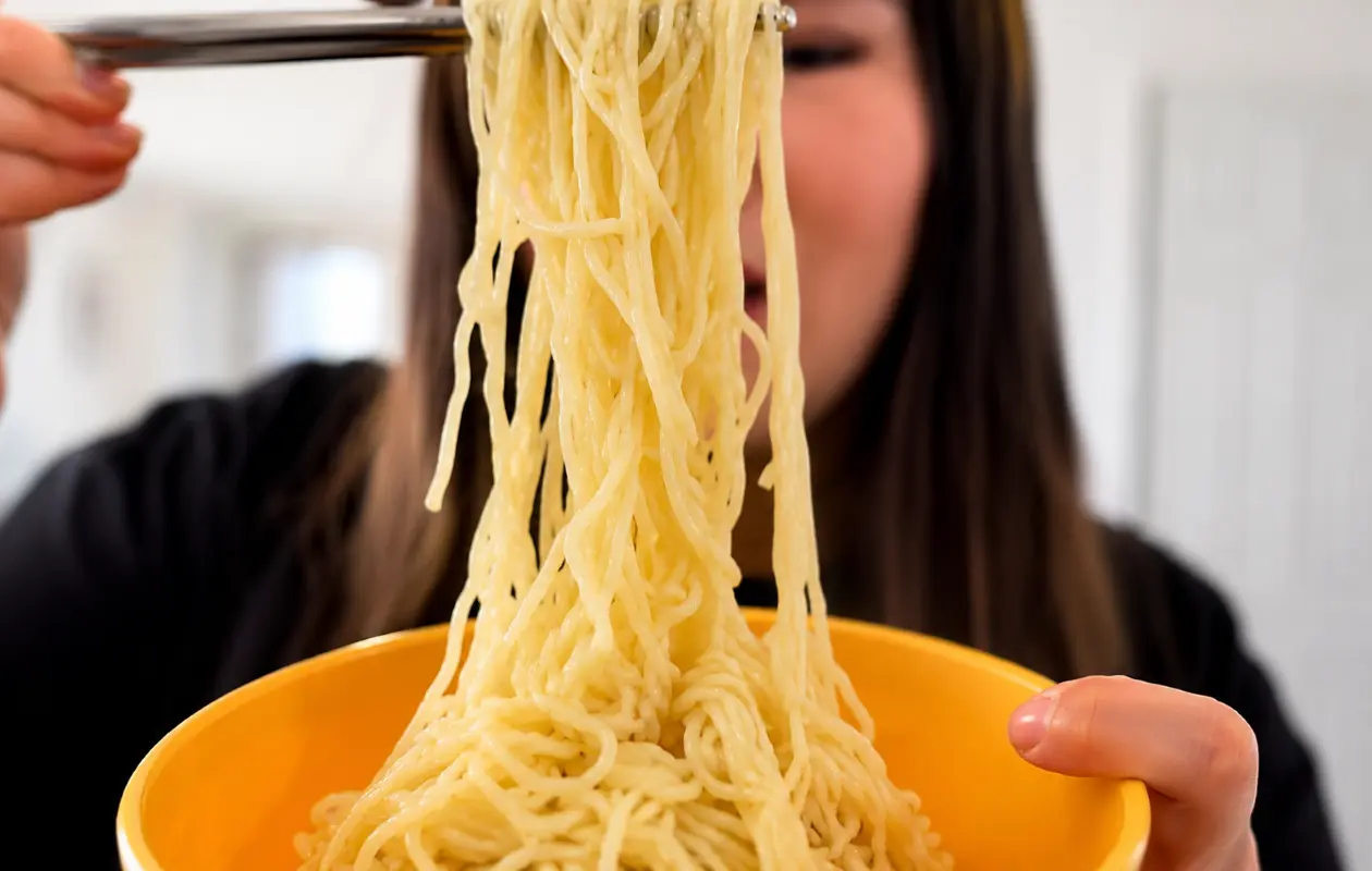 Close-up of glossy, creamy ramen noodles lifted with black chopsticks from a vibrant orange bowl, with the woman softly blurred in the background and the focus on the silky, buttery texture of the noodles.