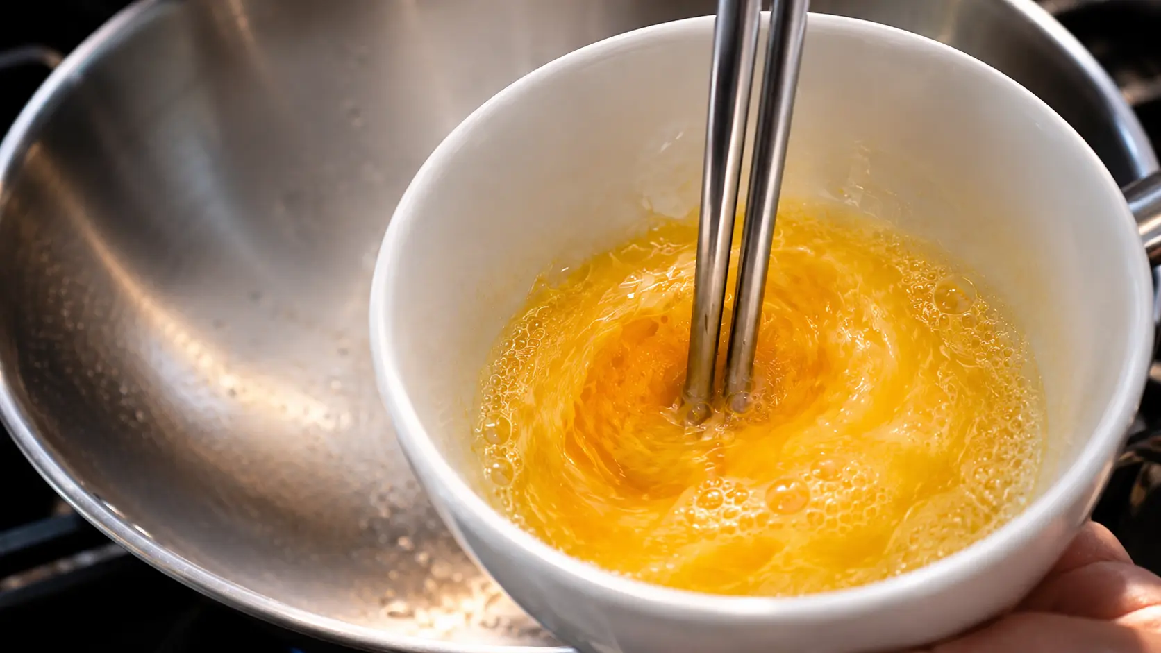 Close-up of a bright yellow egg being whisked with stainless steel chopsticks in a white bowl, with soft bubbles forming on the surface and a blurred stovetop and wok in the background.
