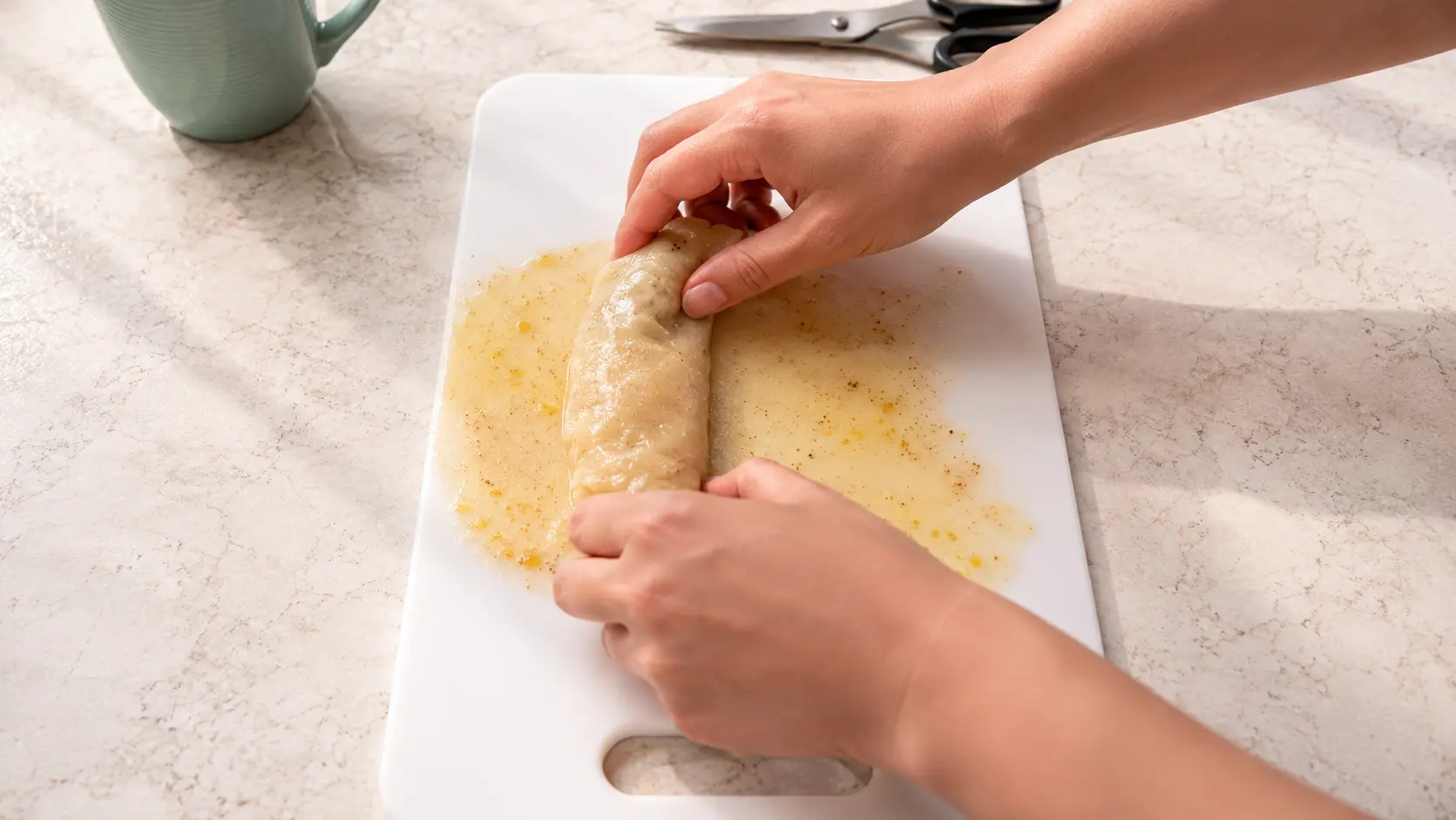 A professional food photography-style close-up shows two hands rolling a cinnamon-speckled rice paper sheet into a croissant-like shape on a white cutting board. The rice paper is coated in a glossy custard mixture, creating a soft sheen that catches the warm natural sunlight. The scene is set on a light marble countertop with a muted green ceramic mug and kitchen scissors softly blurred in the background. The image highlights the texture of the rice paper and the hands-on preparation process, creating a clean, inviting, and cozy kitchen atmosphere.