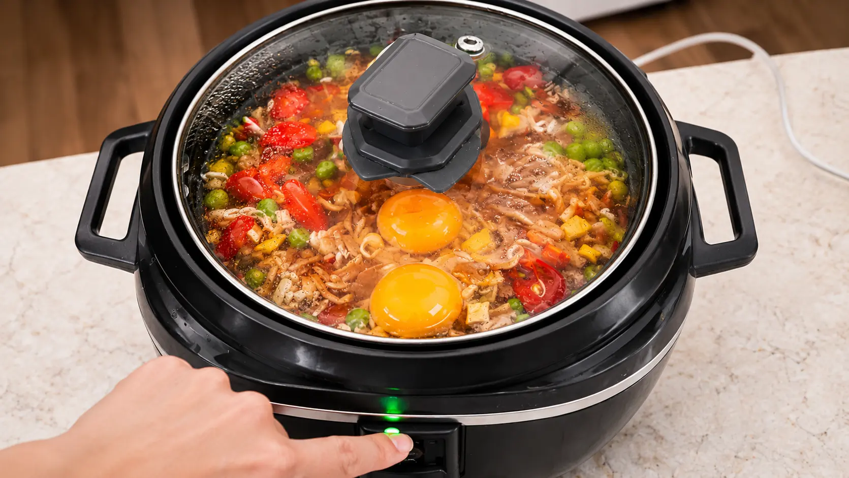 Close-up of a black rice cooker with glass lid as a hand presses the power button, showing rice, vegetables, beef, and two eggs inside on a marble countertop.