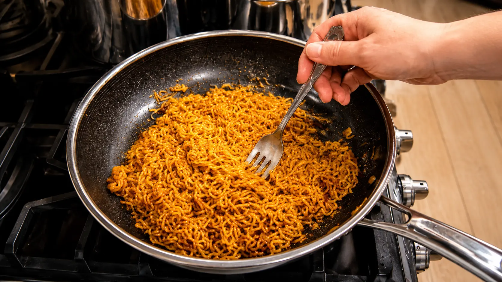Close-up of creamy peanut butter ramen being stirred with a fork in a nonstick skillet on a stovetop, the glossy sauce coating each golden noodle under warm kitchen lighting.