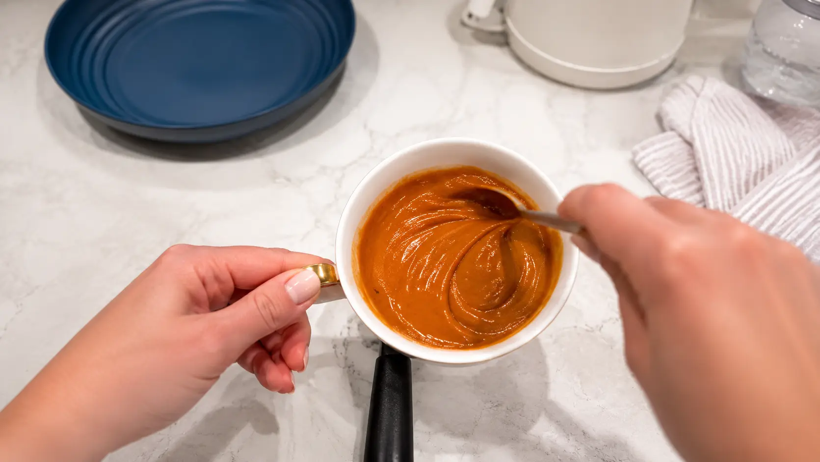 Close-up of creamy peanut butter sauce being stirred in a white bowl with a gold handle, the smooth, glossy mixture swirling richly on a marble countertop.