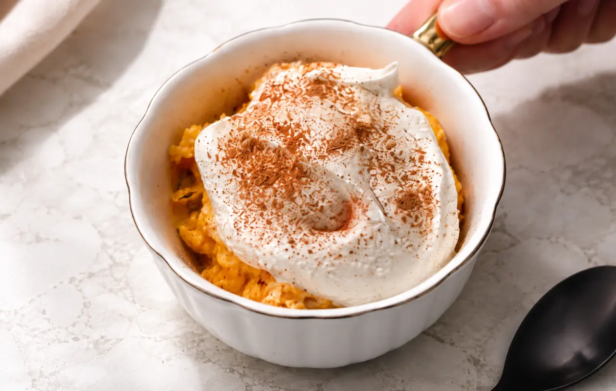 Close-up of a creamy no-bake pumpkin cheesecake in a white scalloped bowl with a gold handle, topped with a thick swirl of Cool Whip and a generous dusting of cinnamon, set on a light marble countertop.