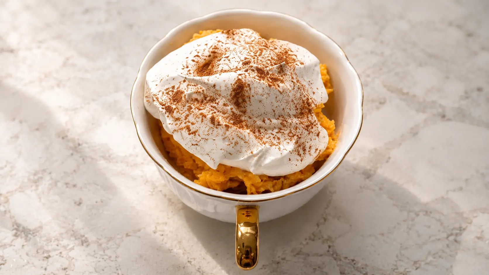 Overhead view of a creamy no-bake pumpkin cheesecake in a white scalloped bowl with a gold handle, topped with fluffy Cool Whip and a dusting of cinnamon, set on a light marble countertop with soft natural lighting.