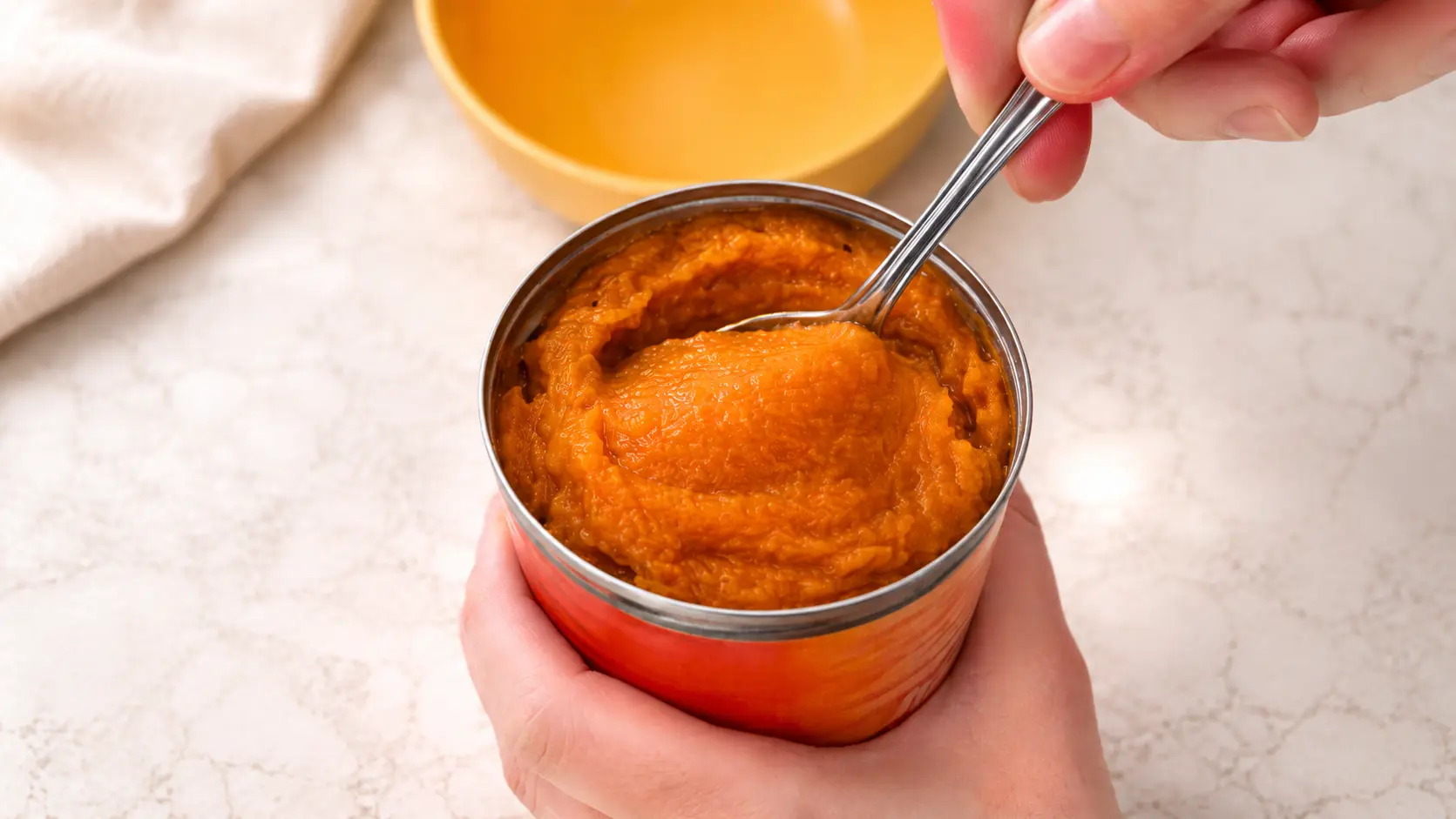 Close-up of hands holding an open can of smooth pumpkin puree while a spoon scoops the thick, vibrant orange mixture, with a yellow bowl blurred in the background on a light marble countertop.