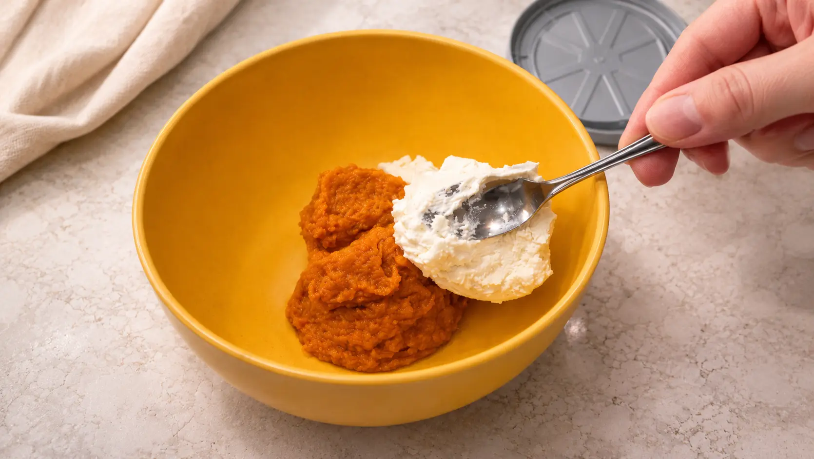 Close-up of pumpkin puree and a scoop of cream cheese in a bright yellow mixing bowl on a light marble countertop, with a hand holding a spoon adding the cream cheese under soft, even lighting.